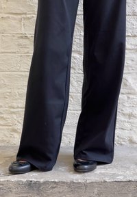 Pantaloni negri cu croi larg, dintr-un material neted, asortați cu pantofi plat negri cu un accent sub formă de fundiță. Fundalul este un perete texturat alb.