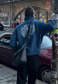 Μπλε φλις μπλούζα με κυκλικό λογότυπο στην πλάτη, φορεμένη με μαύρο παντελόνι και μαύρη τσάντα ώμου, στέκει δίπλα σε ένα αυτοκίνητο.