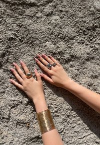 Dwie dłonie z długimi, jasnoróżowymi paznokciami prezentujące złoty pierścionek w kształcie słonia, srebrny pierścionek z kwiatowym wzorem oraz grubą, teksturowaną złotą bransoletkę.