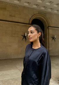 Svart silkesbutton-up plagg med rund krage och lös passform. Bakgrunden har en stenvägg och dekorativa ljuslyktor.