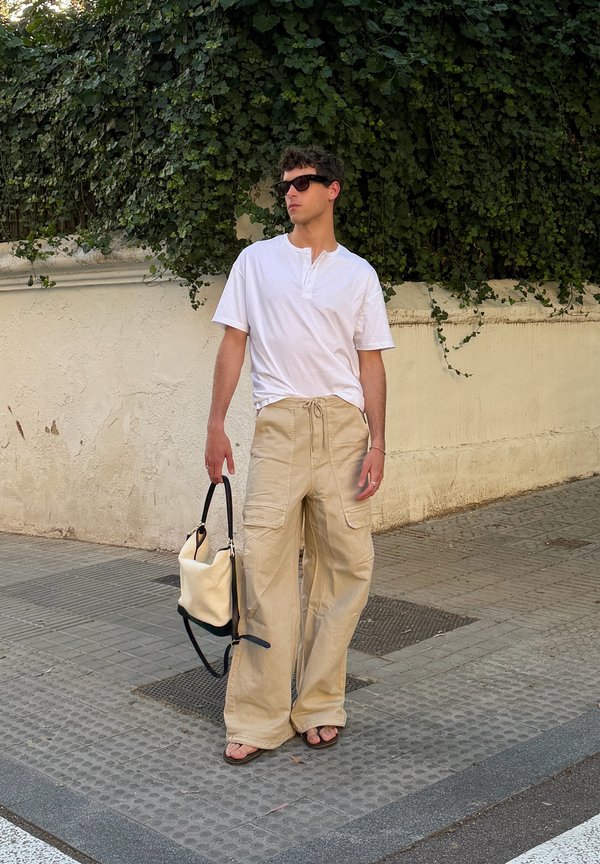 Uomo con occhiali da sole, camicia bianca, pantaloni beige a gamba larga e sandali, in piedi su un marciapiede con una borsa color crema e nera.