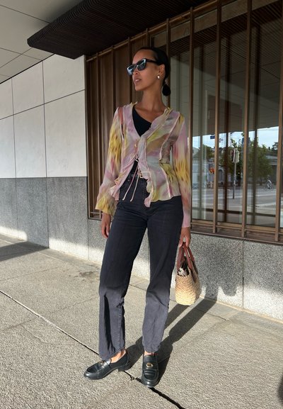 Blusa ligera, multicolor y de manga larga con cintura ajustable, combinada con jeans oscuros y mocasines negros. Sosteniendo una bolsa de mimbre.