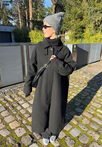 Abrigo de lana negro con un cuello alto, que presenta botones grandes y mangas anchas. Combinado con un gorro de punto gris y un bolso cruzado negro.
