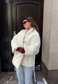 Γυναίκα με φαρδιά λευκή φουσκωτή ζακέτα και κόκκινα γάντια στέκεται με σταυρωμένα τα χέρια μπροστά σε σκοτεινή ξύλινη πόρτα, φορώντας γυαλιά ηλίου.