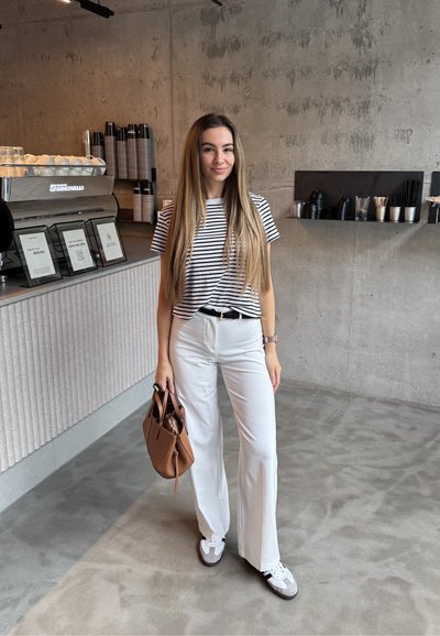 Camiseta a rayas, pantalones anchos blancos y zapatillas grises. Sosteniendo un bolso marrón. Cafetería moderna con paredes y mostrador de hormigón.
