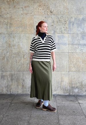 Suéter de rayas blanco y negro de manga corta, falda larga verde oliva, calcetines azules con estampado, zapatos marrones con suela gruesa, de pie contra una pared texturizada.