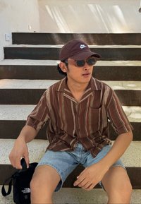Uomo seduto sui gradini, indossando una camicia a maniche corte a righe marroni, shorts di jeans, occhiali da sole e un cappellino scuro. Borsa a tracolla nera accanto.