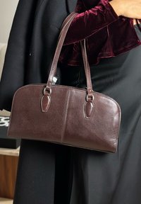 Sac à main en cuir marron de forme arrondie, avec deux poignées supérieures et des accessoires en métal argenté. Surface texturée avec coutures visibles et accents.