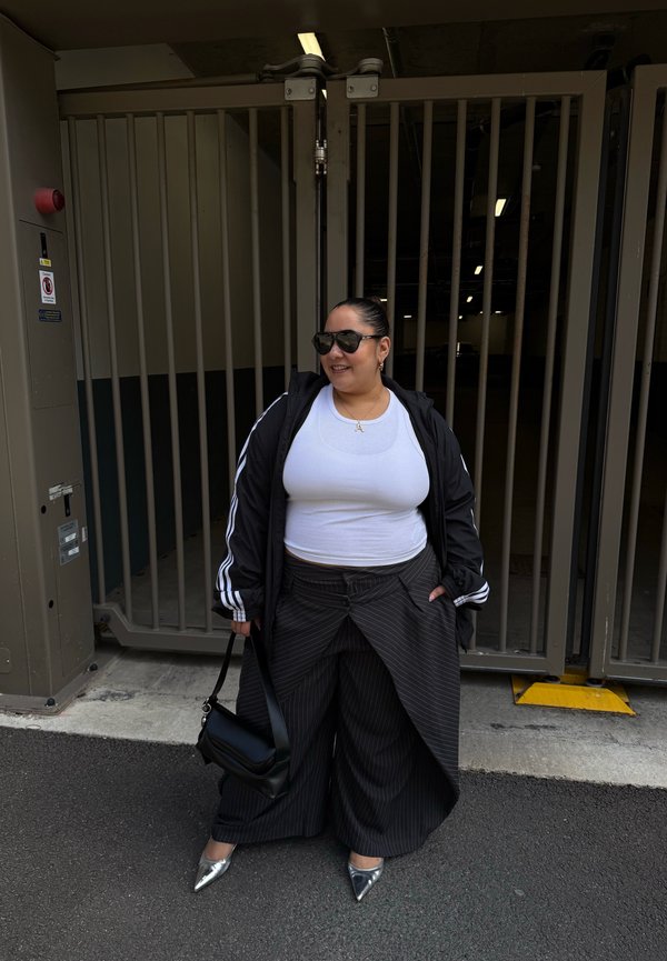 Frau trägt weißes Top, schwarze Jacke, weite Nadelstreifenhose, silberne Schuhe und Sonnenbrille und steht auf dem Bürgersteig vor einem geschlossenen grauen Metalltor.