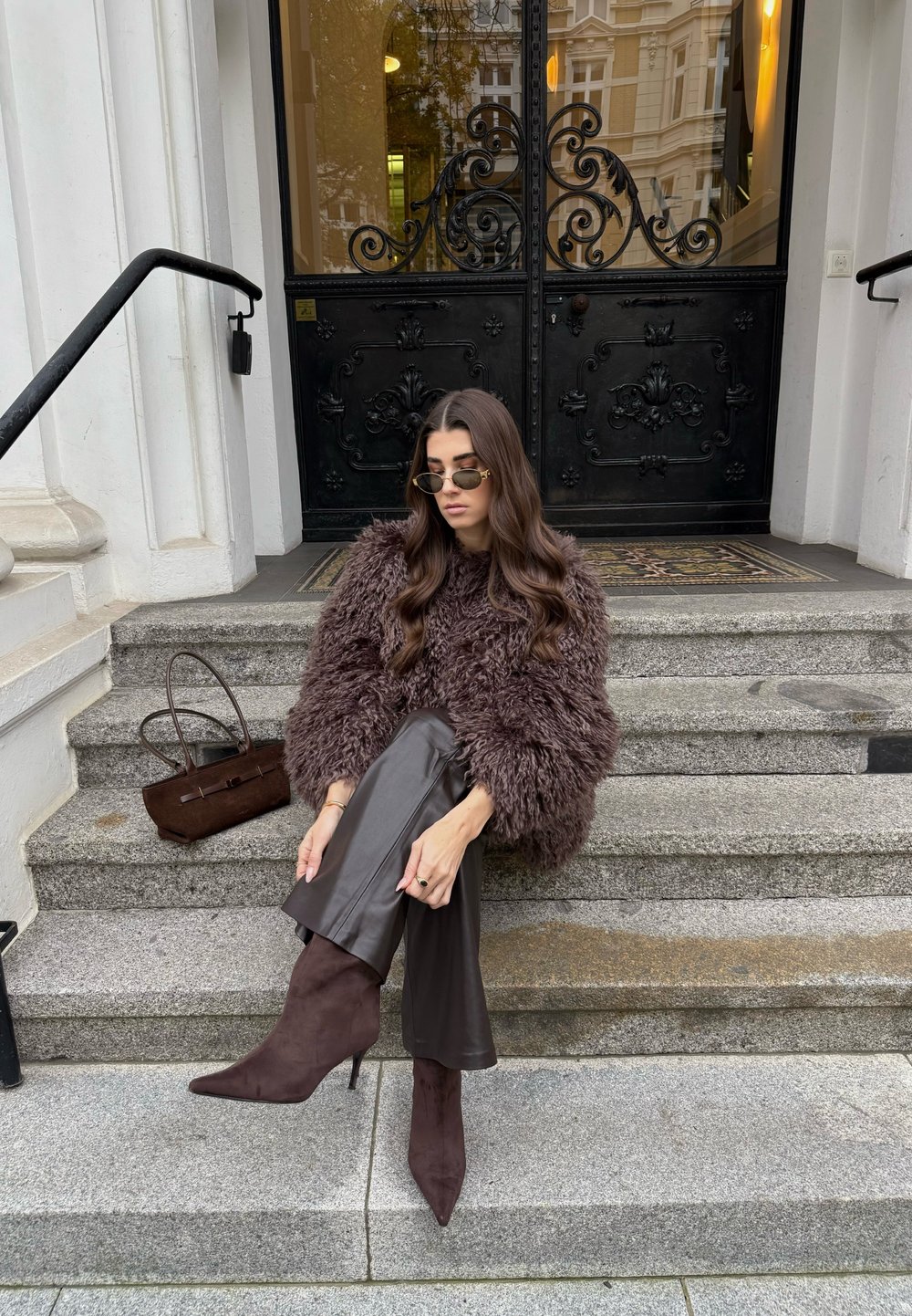 Haină maro de blană artificială, pantaloni mulați din piele maro, cizme maro din piele întoarsă cu toc înalt, ochelari de soare rotunzi și o geantă mică maro pe treptele de piatră.
