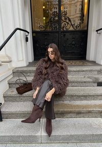 Haină maro de blană artificială, pantaloni mulați din piele maro, cizme maro din piele întoarsă cu toc înalt, ochelari de soare rotunzi și o geantă mică maro pe treptele de piatră.