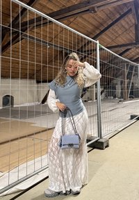 Žena s kučeravými vlasmi v modrom topu a bielych čipkovaných sukni, držiaca malú modrú kabelku, stojaca proti kovovému plotu vnútorných priestorov pod drevenou strechou.
