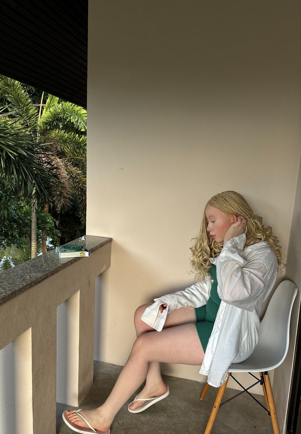 Λευκό πουκάμισο με μακρύ μανίκι πάνω από ένα πράσινο φόρεμα, καθισμένο σε μια ανοιχτόχρωμη ξύλινη καρέκλα. Ανοιχτόχρωμο δέρμα, σγουρά ξανθά μαλλιά. Μαρμάρινη βεράντα με ψηλές παλμίνες ορατές.