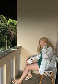 Λευκό πουκάμισο με μακρύ μανίκι πάνω από ένα πράσινο φόρεμα, καθισμένο σε μια ανοιχτόχρωμη ξύλινη καρέκλα. Ανοιχτόχρωμο δέρμα, σγουρά ξανθά μαλλιά. Μαρμάρινη βεράντα με ψηλές παλμίνες ορατές.