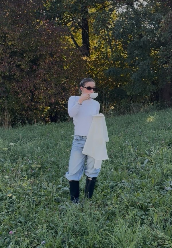Une femme portant des lunettes de soleil sirote une tasse de thé dans une clairière herbeuse, tenant un tissu couleur crème sur fond d’arbres.