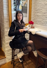 Jonge vrouw in een zwarte jas en rok zit op een kruk bij de balie van een café, met een koffiekopje in de hand en een taart en rode rozen vlakbij.