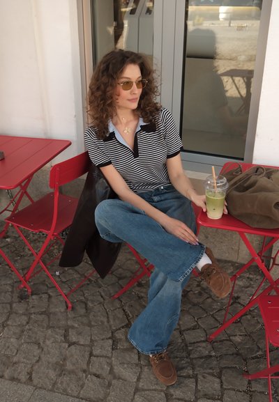 Mujer con cabello rizado y gafas de sol sentada en una mesa roja de un café al aire libre, sosteniendo una bebida verde con hielo, lleva una camisa de rayas, vaqueros y zapatos marrones.