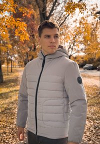 Ανοιχτό γκρι μπουφάν με μόνωση, με μπροστινό φερμουάρ, οριζόντια ραμμένη υφή και στρογγυλή μαύρη επένδυση λογότυπου στο αριστερό μανίκι. Έχει προσαρτημένη κουκούλα.