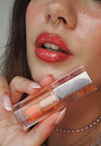 Transparenter Lipgloss in einer klaren Tube mit silberner Kappe, orangefarbenem Farbton und glänzendem Finish. Die Lippen des Models sind in derselben Farbe hervorgehoben.