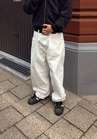 Persona vestida con pantalones blancos holgados y zapatillas negras de pie sobre un pavimento de baldosas cerca de la esquina de un edificio rojo y gris.