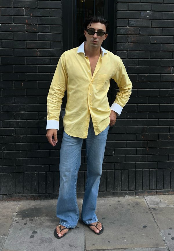 Homme portant une chemise jaune avec des poignets blancs, un jean bleu, des lunettes de soleil noires et des tongs, debout contre un mur en briques noires.