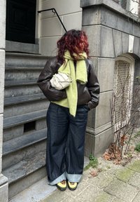 Jaqueta de pele castanha, cachecol verde-lima, calças de ganga azuis oversized e sapatos amarelos com padrão de leopardo, segurando uma pequena mala de creme.