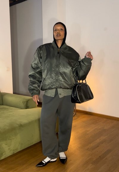 Chaqueta bomber de satén verde, pantalones grises, bolsa negra. Zapatos de punta negra con acentos blancos. Tejido ligero y ajuste relajado.