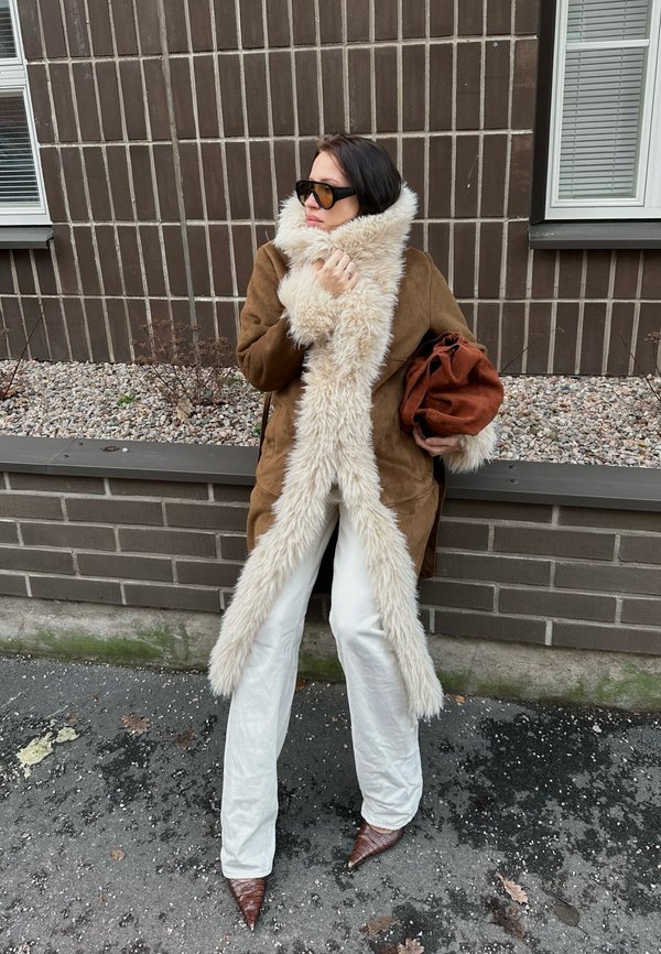 Donna in piedi accanto a un muro di mattoni indossando un cappotto marrone con ampio colletto di pelliccia, pantaloni bianchi, scarpe marroni appuntite, occhiali da sole neri, che tiene una borsa marrone.