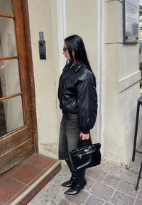 Giubbotto bomber in pelle nera con maniche a sbuffo, abbinato a jeans scuri e stivali neri a punta. Una piccola borsa nera è tenuta in mano.