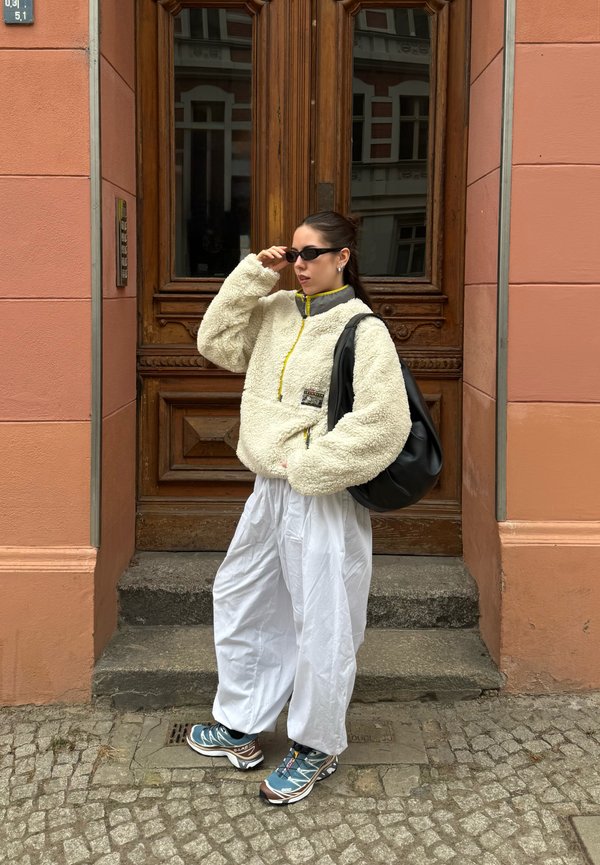 Nainen säätää mustia aurinkolaseja, pukeutuneena vaaleaan fleecetakkiin, valkoisiin väljihin housuihin, sinisiin tennareihin ja kantaa suurta mustaa olkalaukkua puisella ovella.