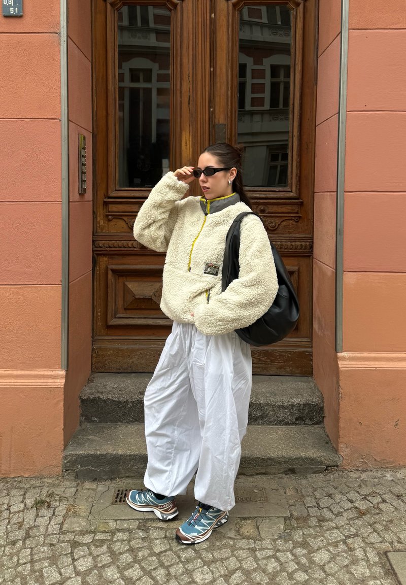 Femme ajustant des lunettes de soleil noires, portant une veste en polaire crème, un pantalon blanc ample, des baskets bleues, et portant un grand sac à bandoulière noir près d'une porte en bois.