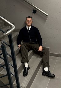 Črn zip-up pulover, bela srajca, črn kravata, olivno zelene hlače, črne čevlje, bele nogavice. Sedi na sivih pločicah s stopnicami in kovinsko ograjo.