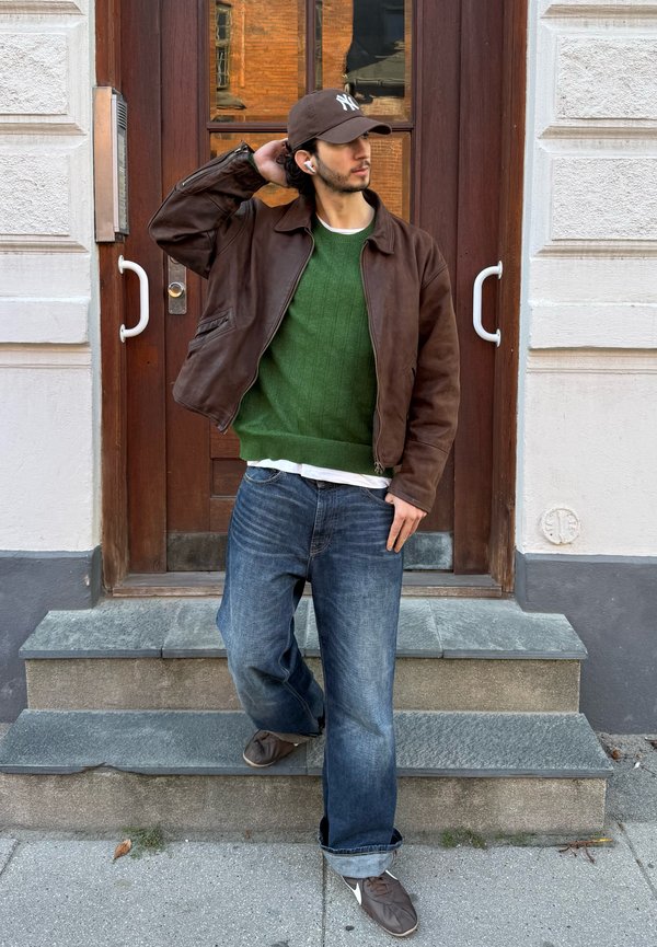 Un hombre con una chaqueta marrón, un jersey verde, vaqueros y una gorra está de pie en unos escalones de piedra, frente a una puerta de madera, ajustándose la gorra.