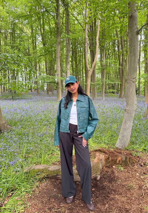Donna con giacca verde, pantaloni neri e cappellino verde che sta su terreno forestale con fiori viola e alberi alti sullo sfondo.