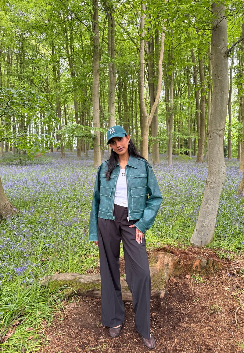 Femme portant une veste verte, un pantalon noir et une casquette verte, debout sur un sol forestier avec des fleurs violettes et de grands arbres en arrière-plan.