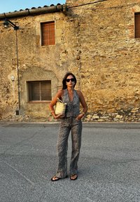 Dvojdielny outfit s leopardím potlačou, ktorý obsahuje bezrukávový top a rozšírené nohavice, doplnený tkanou taškou. Pozadie tvorí kamenná stena.