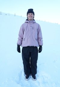Pink insulated jacket, black trousers, and gloves, standing in the snow. Notable details include zippers and pockets on both the jacket and trousers.