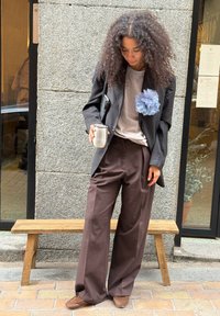 Sort stribede blazer over en lys grå skjorte, brune brede bukser, holder en sølvkop; blå stofblomst på blazeren.