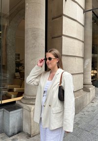 Svijetložuti laneni sako s dva gumba, uparen s bijelom majicom i širokim hlačama. Dodatci uključuju sunčane naočale s uzorkom i malu torbicu.