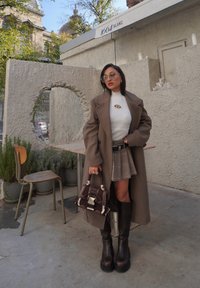 Manteau en laine marron, pull en tricot blanc à côtes, jupe à carreaux, bottes marron montant aux genoux et petit sac à main. Lunettes de soleil transparentes et bijoux minimalistes. Cadre urbain.