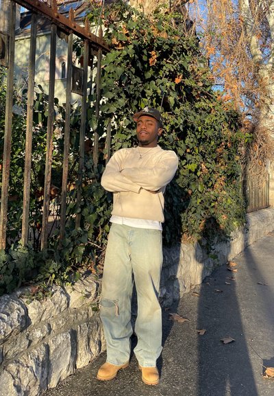 Una persona está de pie con los brazos cruzados, vistiendo un suéter beige claro, una camisa blanca y unos jeans azul claro, complementados con botas marrones.