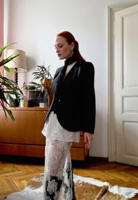 Svart skreddersydd blazer med lange ermer og avrundet kant, lagd over en gjennomsiktig hvit topp og en blonderkjørt med blomsterdekor.