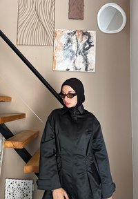 Μαύρο σατέν παλτό με ψηλό γιακά, πλαϊνές τσέπες και διαμορφωμένα μανίκια. Το φόντο περιλαμβάνει έργα τέχνης και ξύλινες σκάλες.