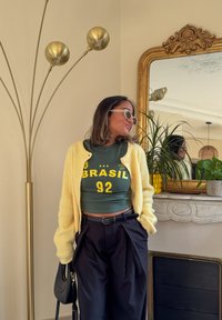 Yellow knit cardigan over a green top with "BRASIL 92" in yellow, paired with black trousers; background features a mirror and houseplants.