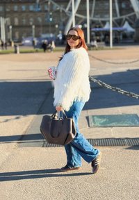 Femme portant une veste blanche à poils longs et un jean bleu, tenant un grand sac à main noir et une canette, debout en plein air par une journée ensoleillée.