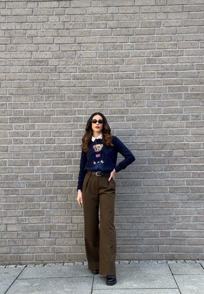 Mujer con cabello largo que lleva gafas de sol, suéter azul marino con diseño de oso, cuello blanco, pantalones marrones de pierna ancha y zapatos negros, de pie contra una pared de ladrillos.