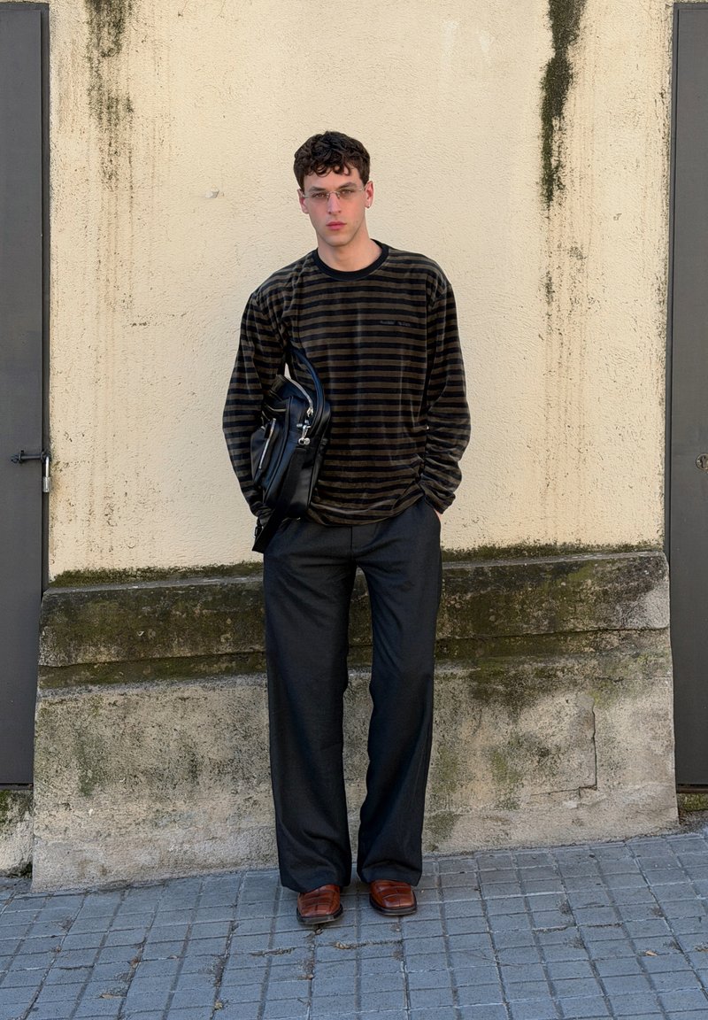 Jeune homme aux cheveux courts et bouclés portant des lunettes transparentes, une chemise à manches longues rayée, un pantalon large noir, des chaussures marron, et portant un sac noir, debout.