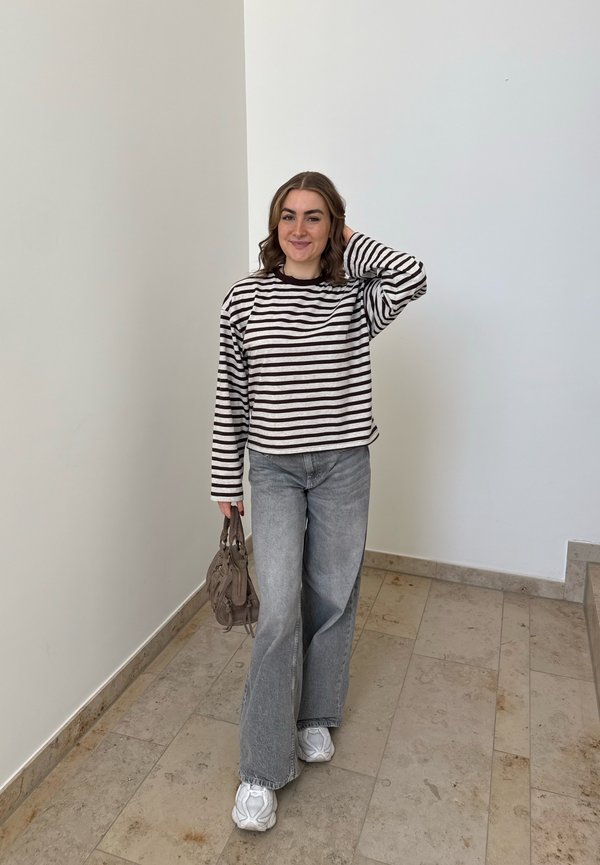 Glimlachende vrouw in gestreept shirt met lange mouwen en brede grijze jeans staat binnen, ze houdt een bruine handtas vast en raakt met één hand haar haar aan.