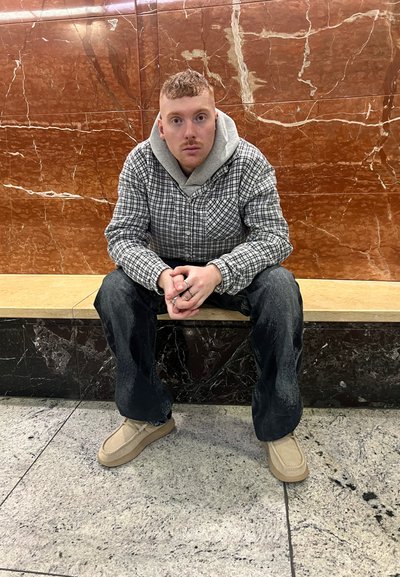 Camisa de franela a cuadros sobre un hoodie gris, jeans negros y zapatos beige. Sentado en un banco de piedra contra un fondo de mármol con patrón.