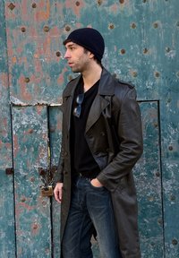 Άνδρας με μαύρο σκουφί, δερμάτινο παλτό και τζιν στέκεται με το χέρι στην τσέπη μπροστά από μια φθαρμένη τιρκουάζ μεταλλική πόρτα με σκουριασμένες βίδες.